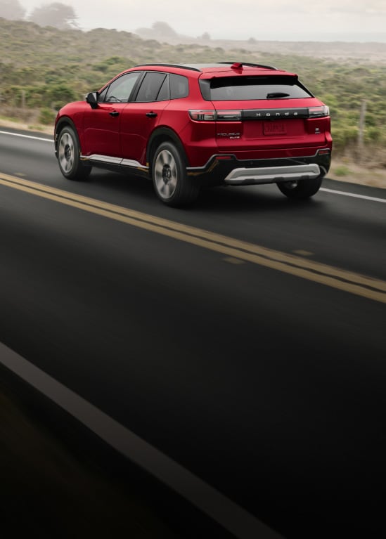 2026 Honda Prologue Elite in Scarlet Red Metallic rear view, driving on a scenic coastal highway (All-Electric SUV).