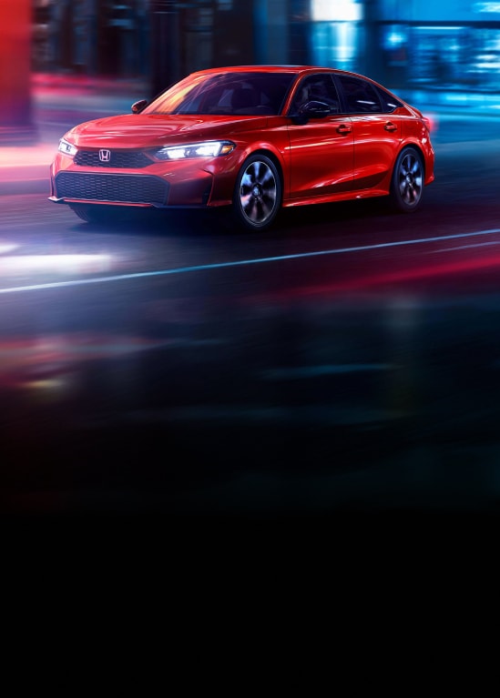 2027 Honda Civic Sedan Sport Touring Hybrid in Rallye Red front driving on a city street at night (Compact Sedan).