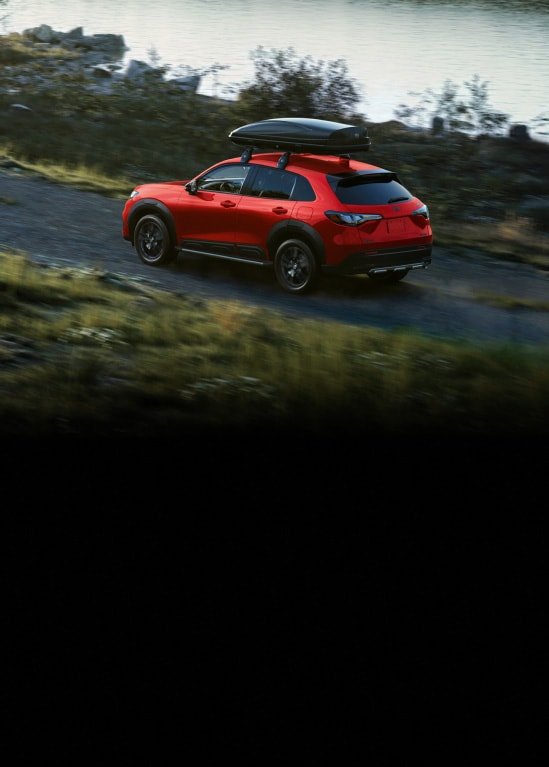2027 Honda HR-V Sport shown in Milano Red with Honda Genuine Accessories, rear view, driving along a scenic lakeside highway (Subcompact Crossover SUV).