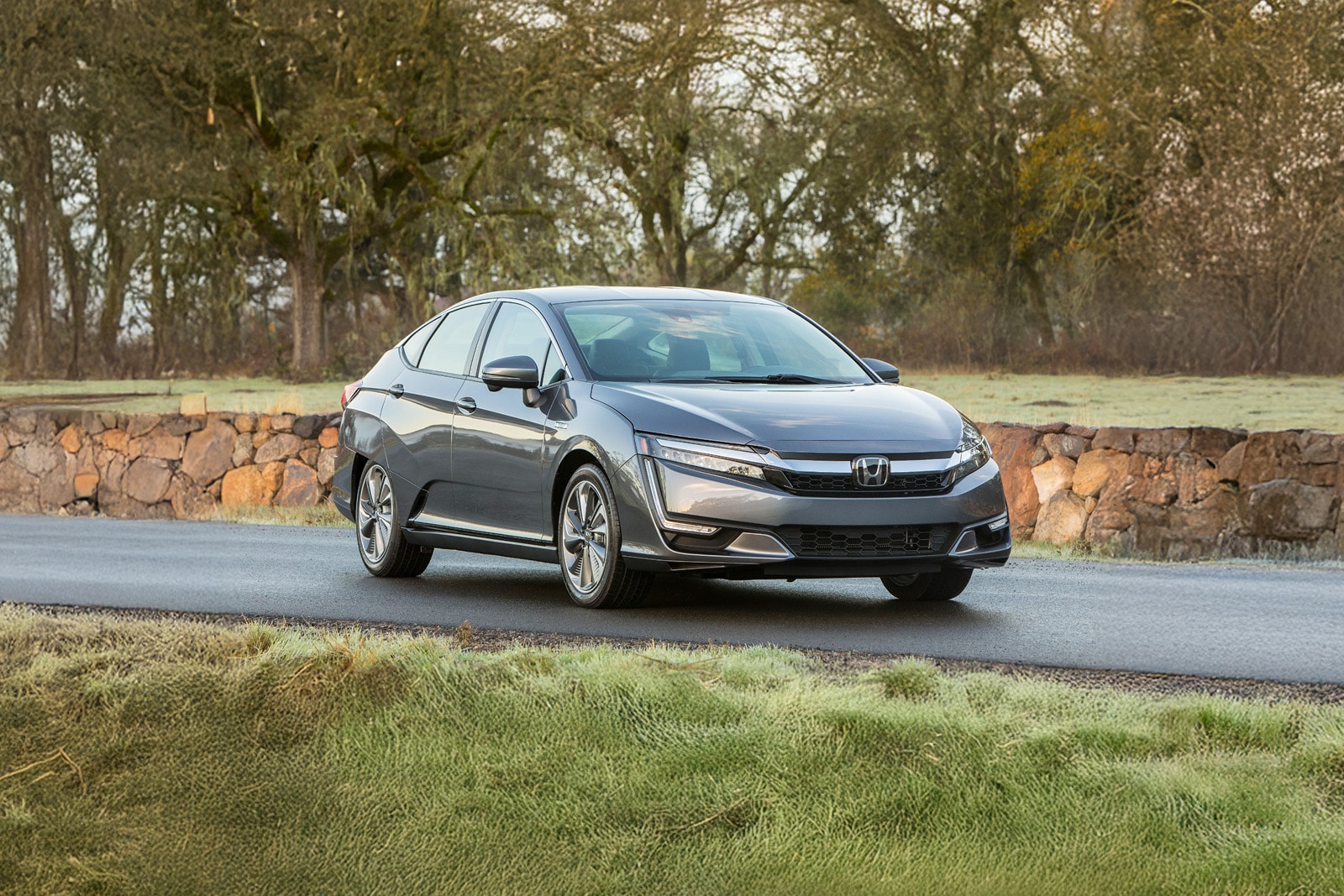 2020 Honda Clarity Plug-In Hybrid in Modern Steel Metallic, front view, driving on a paved road past a stone wall and green grass.